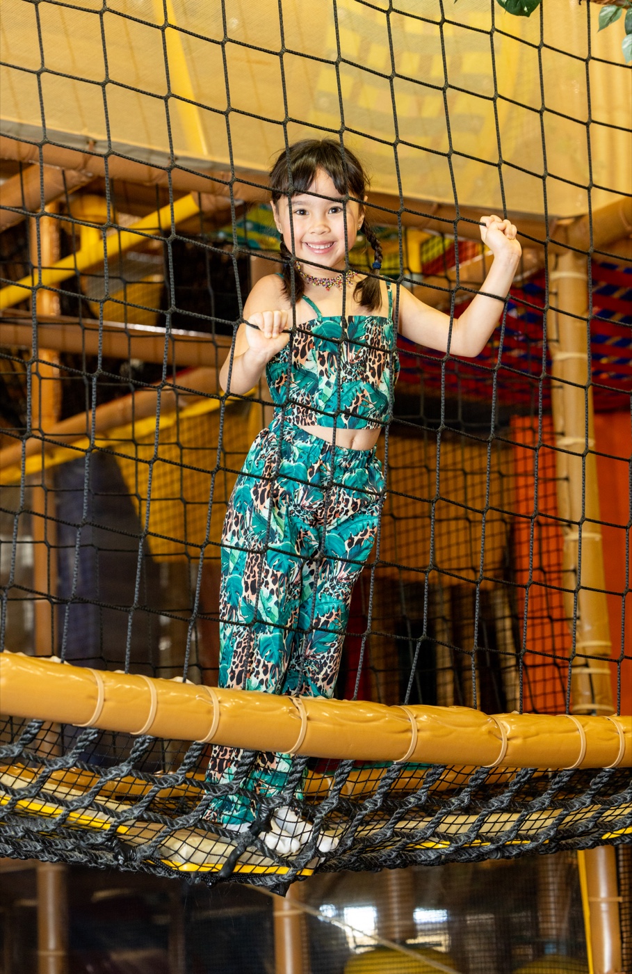 A smiling girl in a jungle-print outfit standing on the netting inside the Safari Run Plano play structure