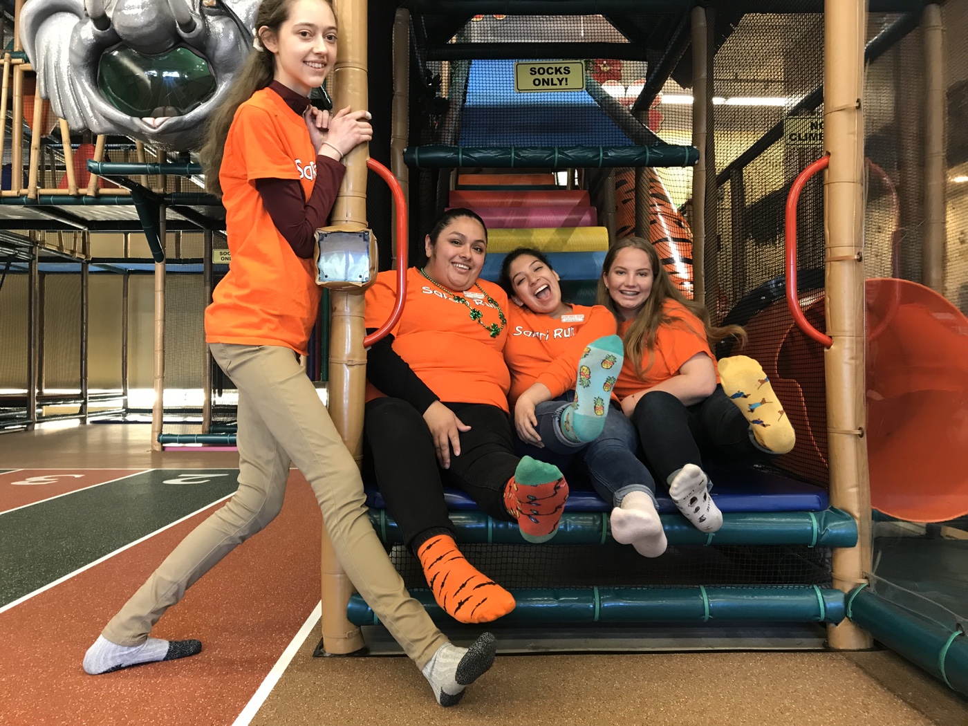 Four Safari Run team members in orange Safari Run t-shirts and animal socks, laughing together inside the play structure