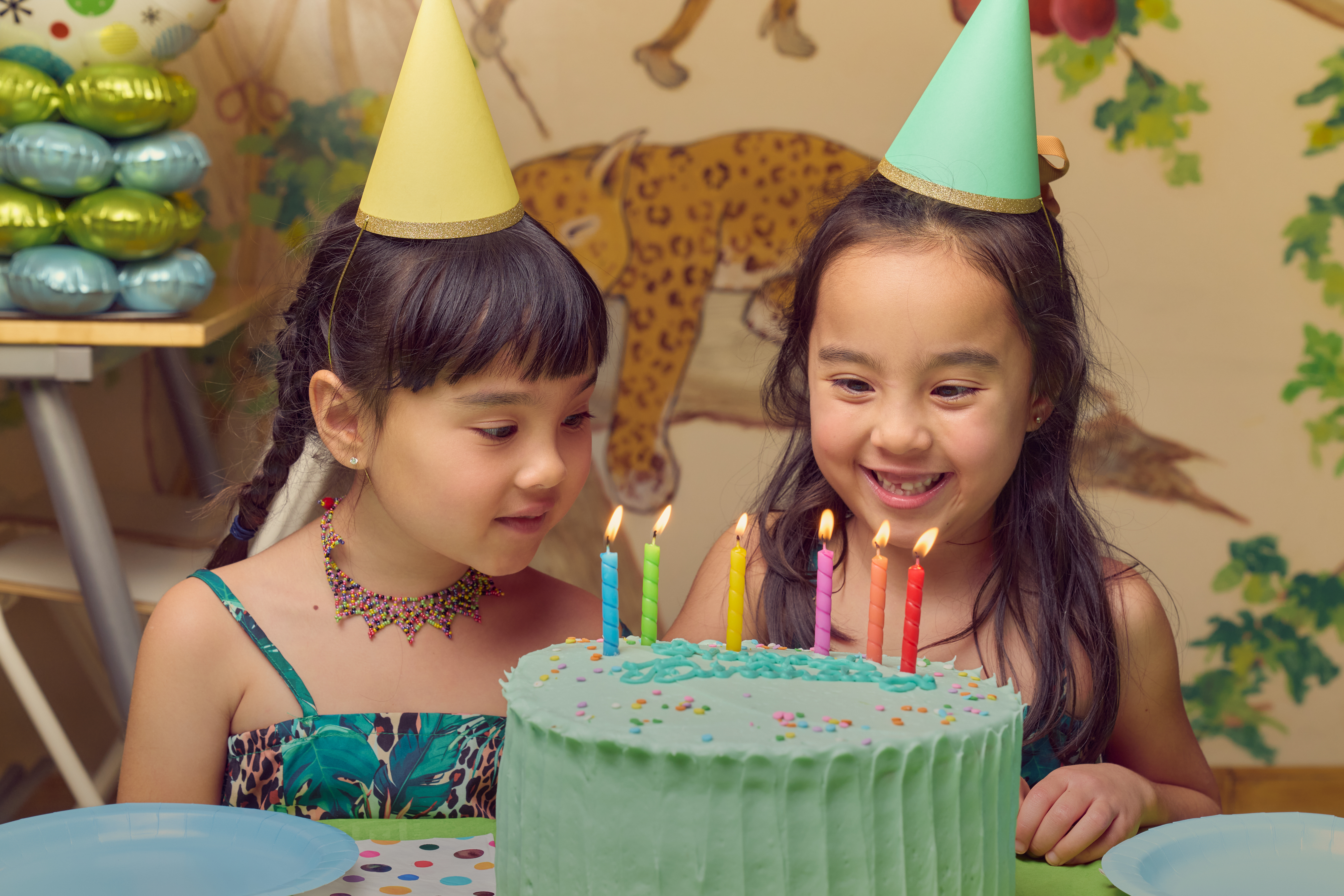 Two girls in party hats blowing out candles on a green birthday cake at Safari Run